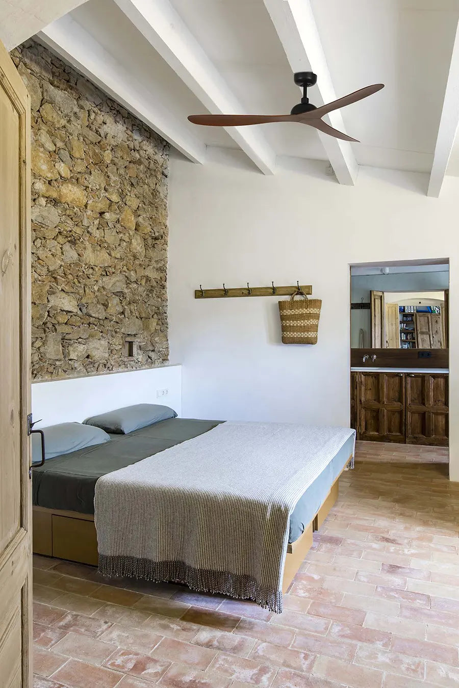 Ventilador de techo Gotland en acabado negro con aspas en tono nogal, instalado en un dormitorio de techo abuhardillado con vigas blancas y pared de piedra
